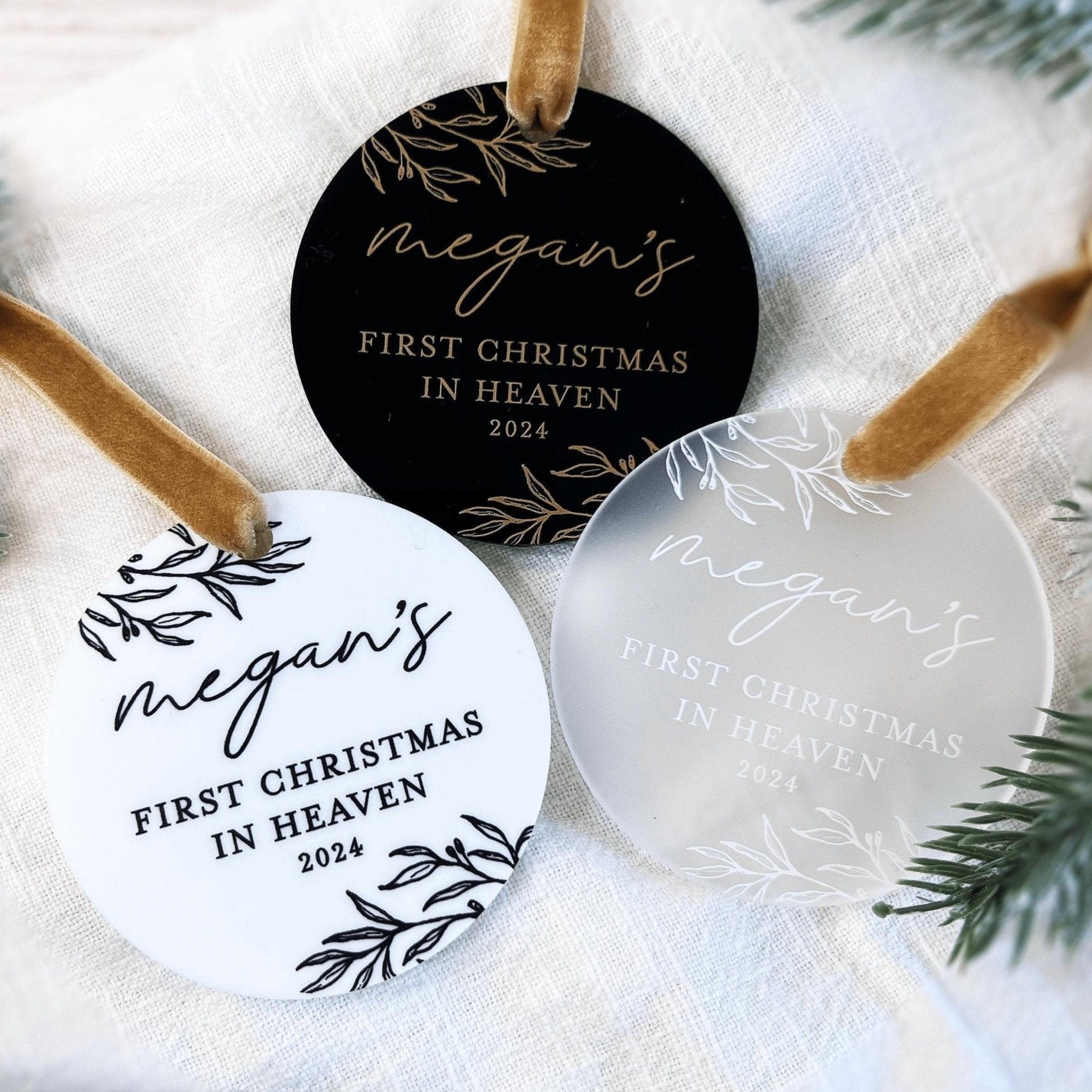Three round acrylic ornaments in black, white, and frosted finishes with personalized names and "First Christmas in Heaven 2024" text with botanical leaf designs.