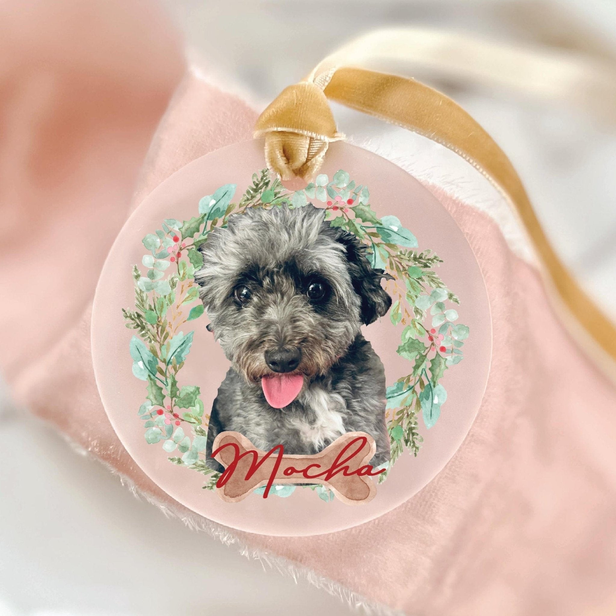 Personalized pet photo ornament with schnauzer portrait surrounded by green floral wreath and red "Mabel" text, clear acrylic with gold ribbon