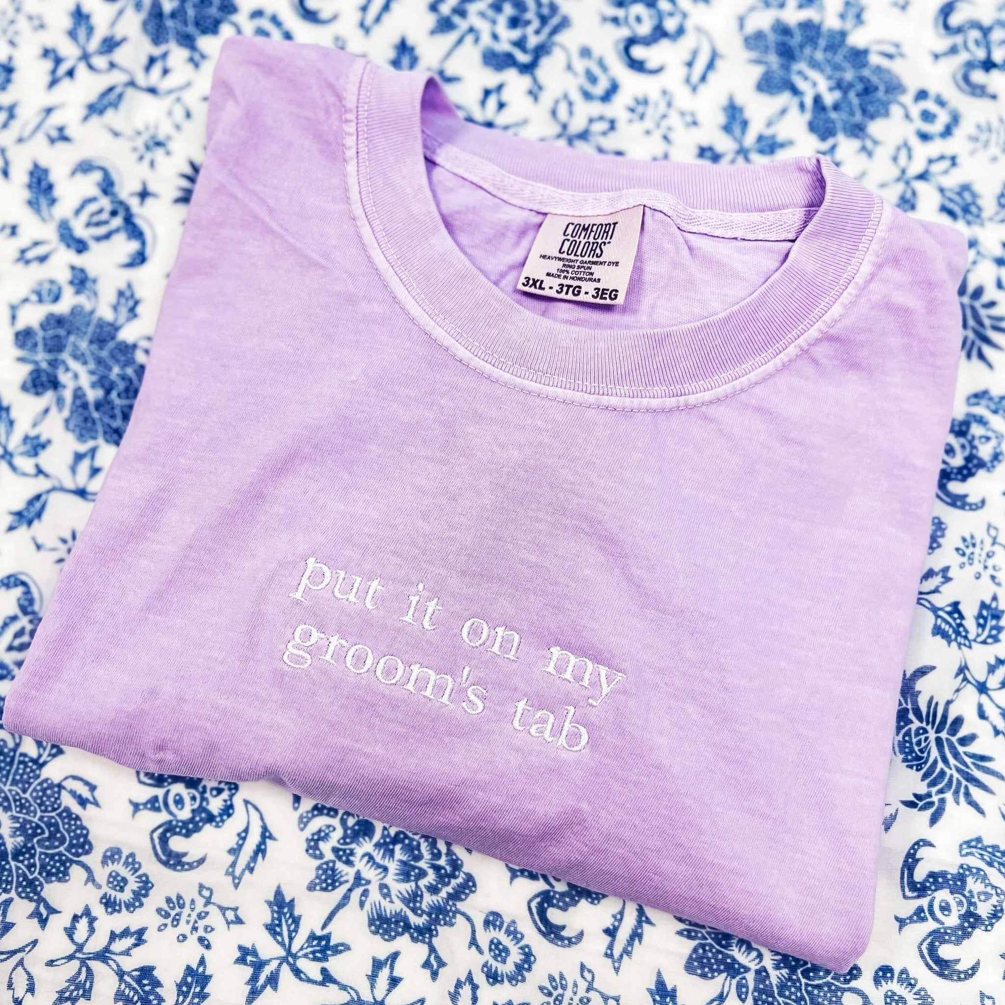 Lavender t-shirt with white embroidered text reading "put it on my groom's tab" on blue floral background.