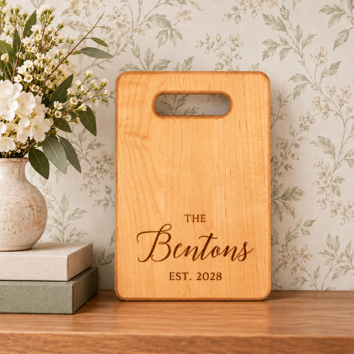 Personalized maple wood cutting board with engraved Benton family name and established year 2028, displayed upright with floral decor.