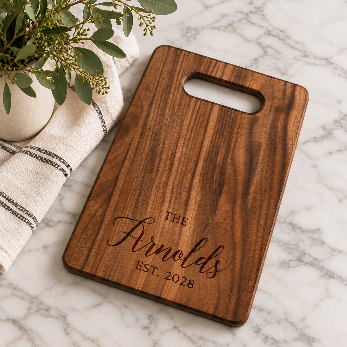 Custom walnut cutting board with engraved Arnold family name and established year 2028, styled on a marble countertop with greenery.