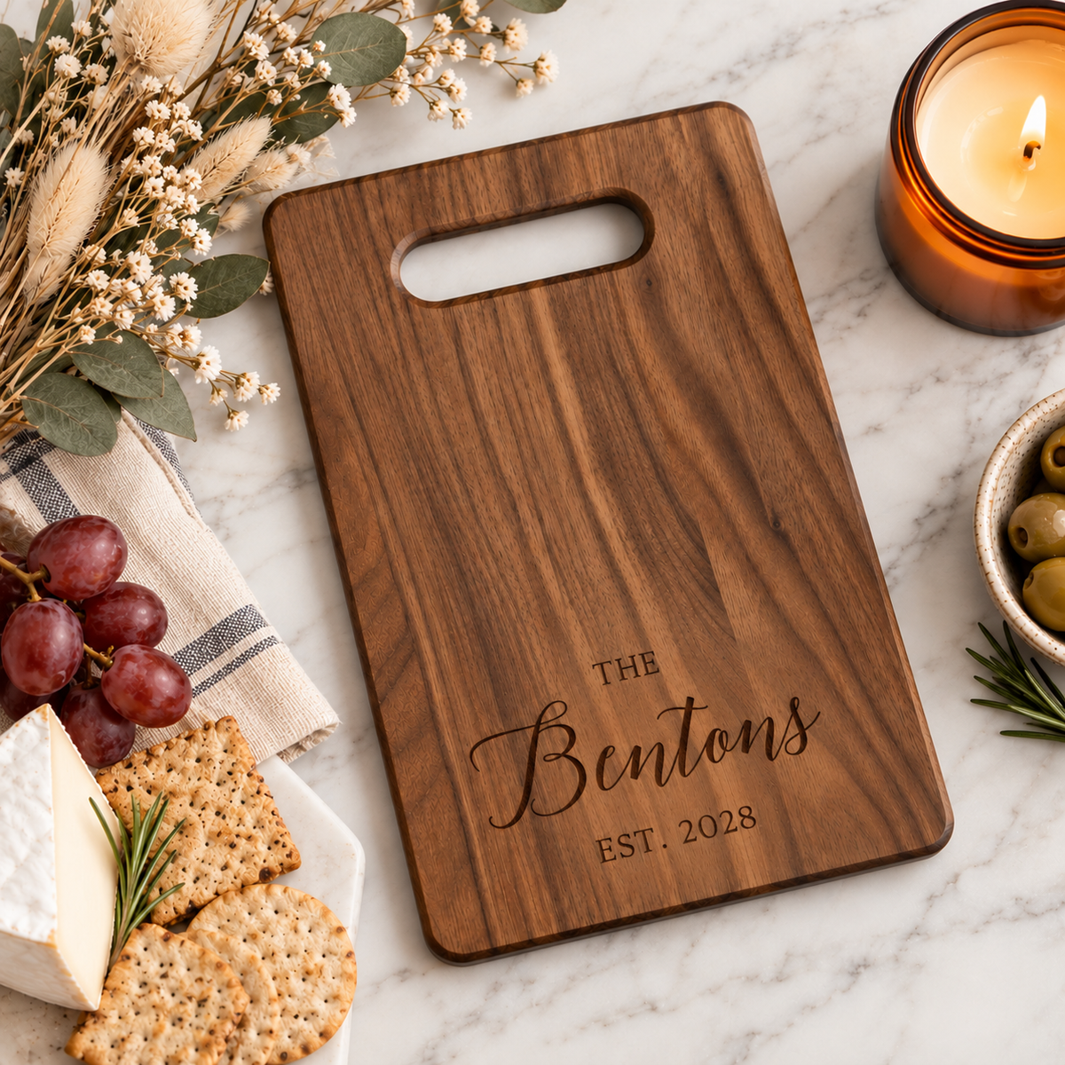 Personalized walnut wood cutting board engraved with Benton family name and established year 2028, styled with cheese, crackers, grapes, and candle.