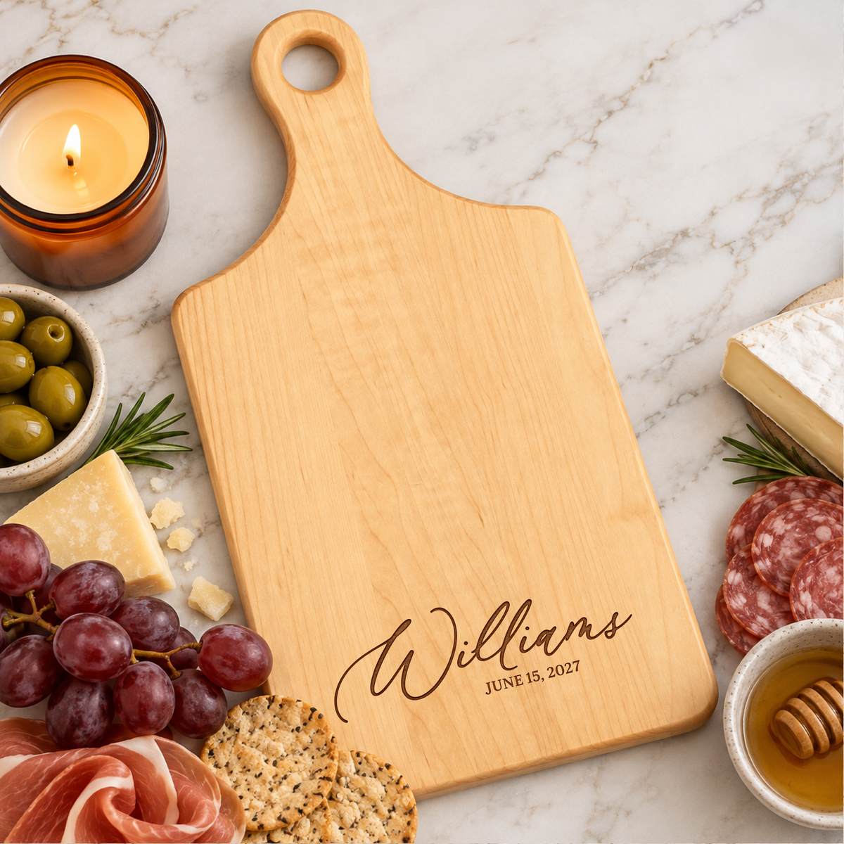 Personalized maple paddle cutting board engraved with Williams and wedding date, styled with cheese, grapes, crackers, and candle on marble countertop.