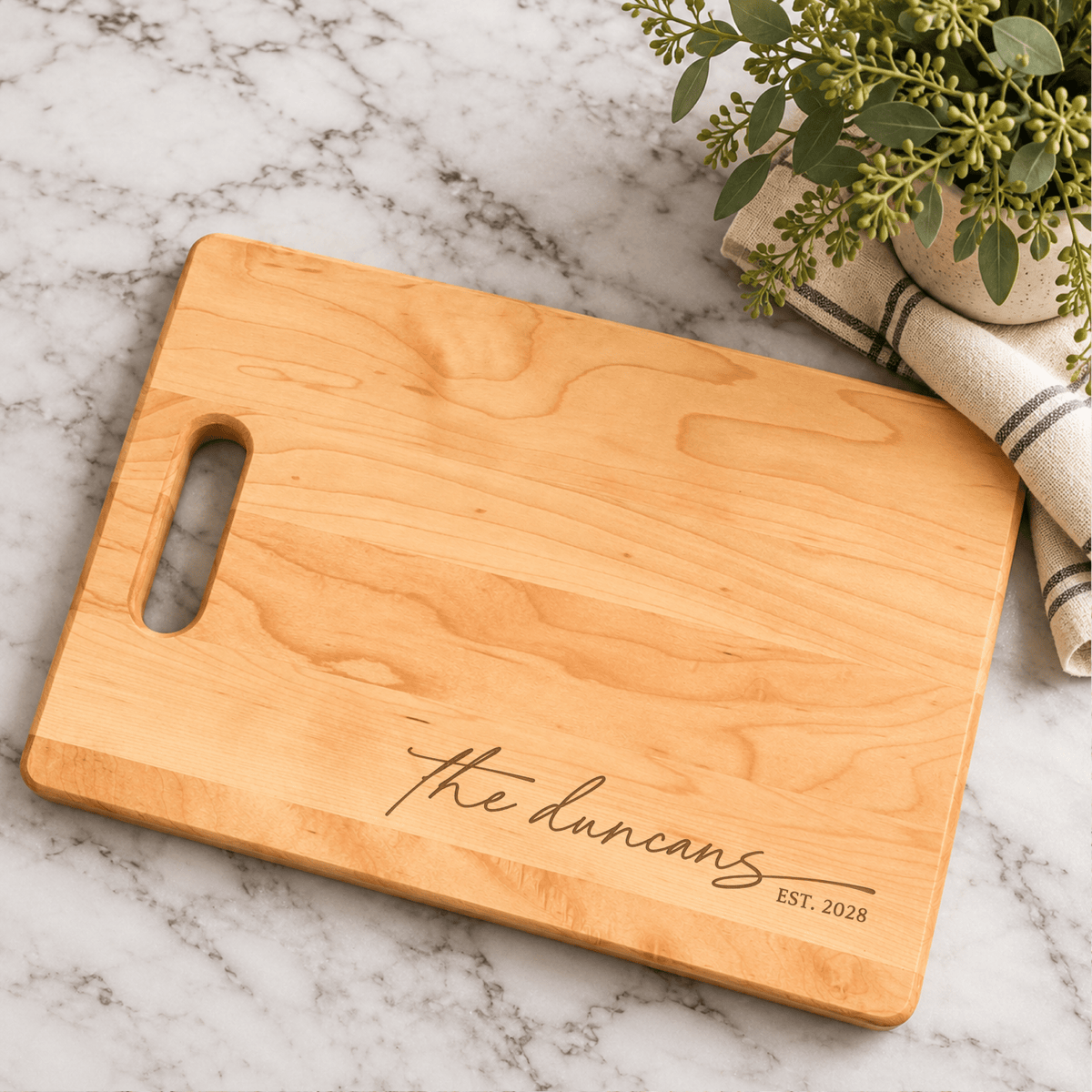Personalized maple cutting board with Duncan family name engraving and established year, styled on marble countertop with greenery and striped kitchen towel.