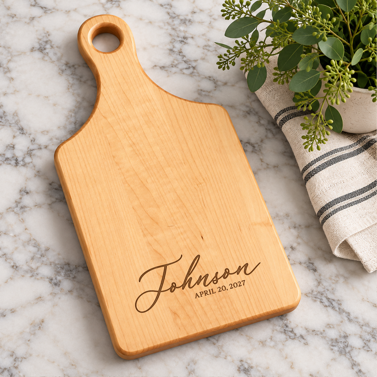 Custom engraved maple wood paddle cutting board featuring Johnson family name and date, displayed on a marble countertop with greenery and kitchen towel.