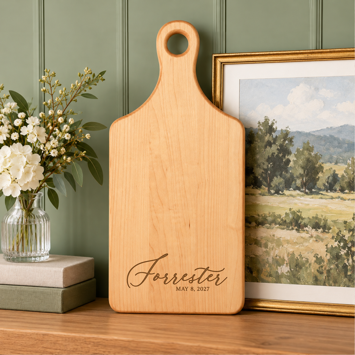 Personalized wooden charcuterie paddle board engraved with Forrester and wedding date, displayed upright with white flowers and framed artwork.