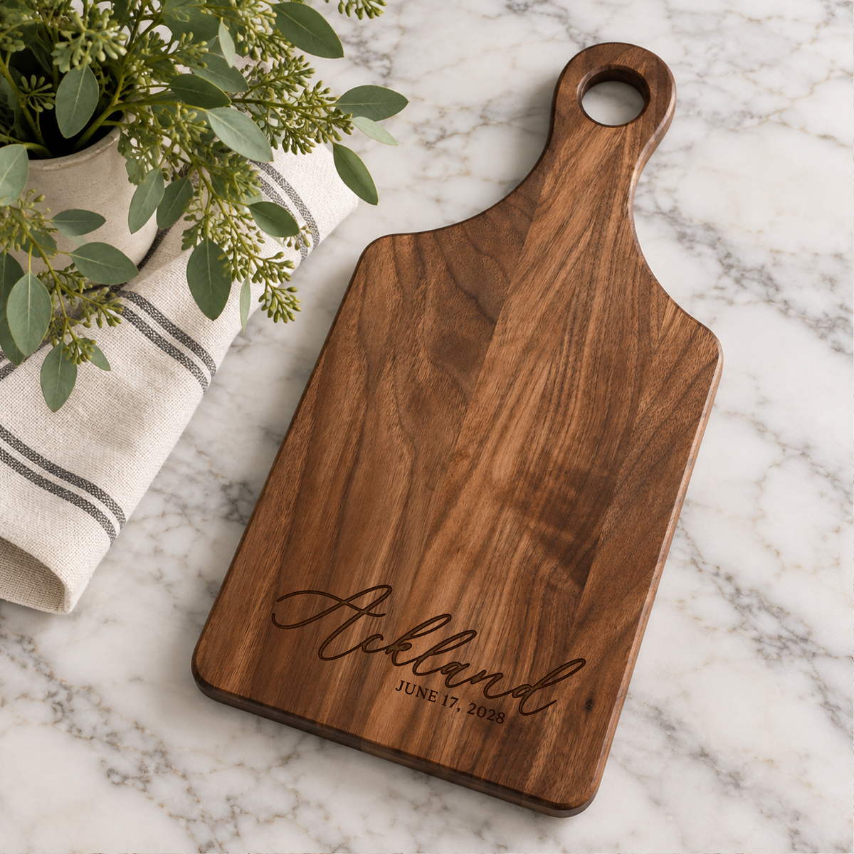Custom walnut paddle cutting board engraved with Ackland and wedding date, styled on a white marble countertop with greenery and striped kitchen towel.