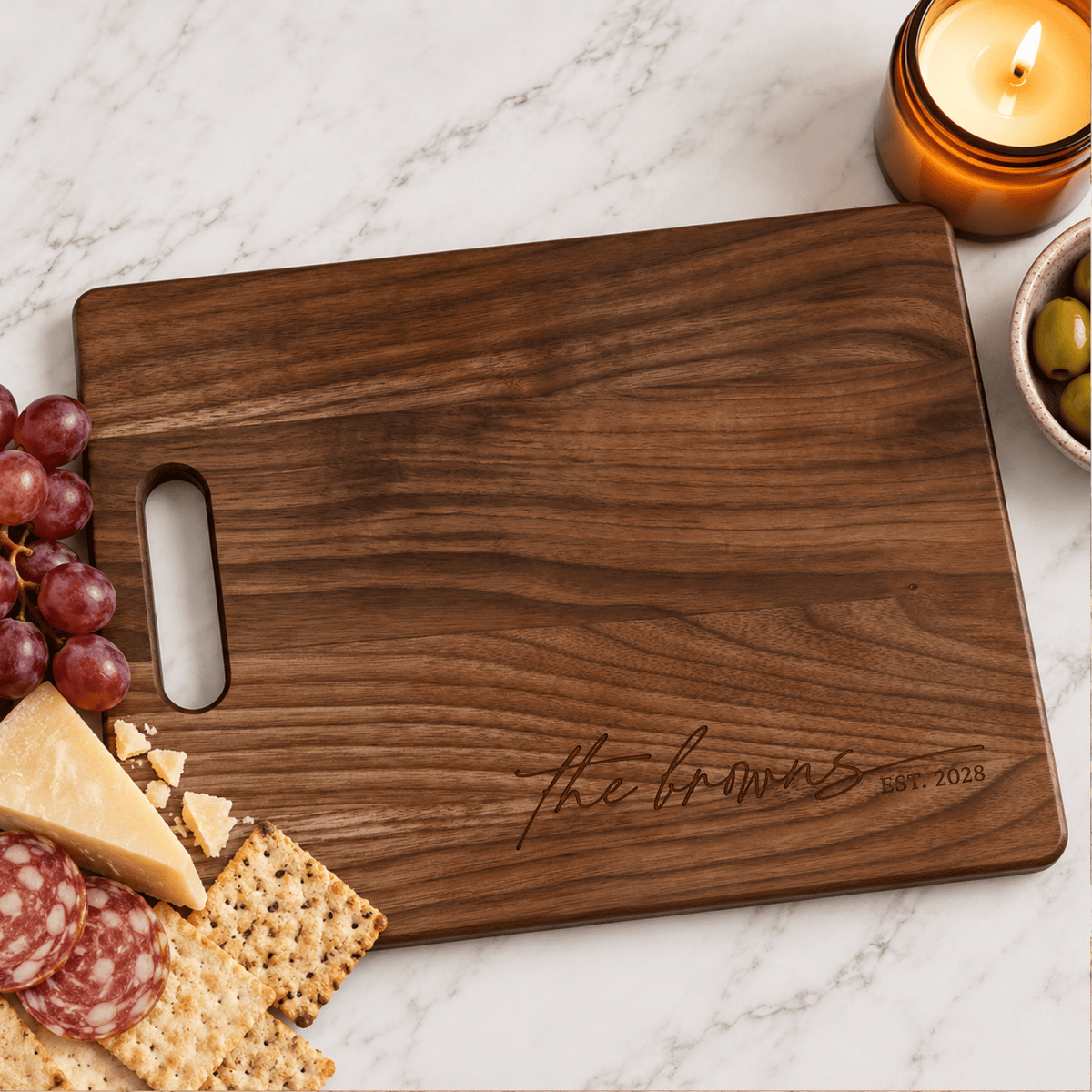 Personalized walnut cutting board featuring Brown family name and established year, styled with grapes, cheese, crackers, olives, and candle on marble.