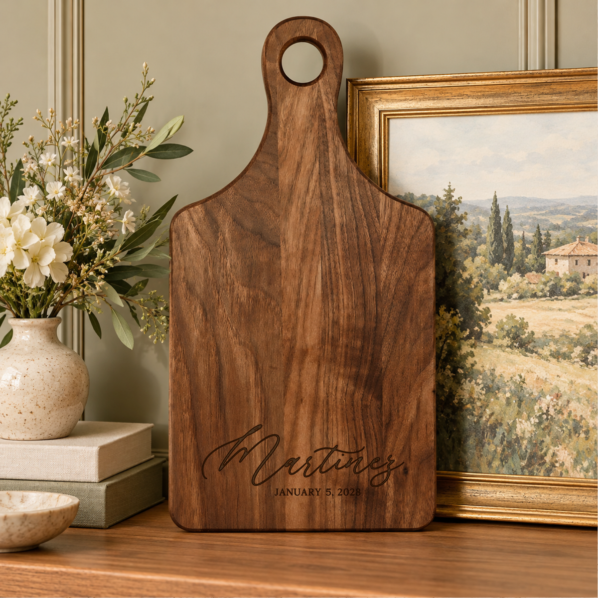 Personalized walnut wood serving board featuring Martinez surname and engraved date, displayed upright with floral arrangement and framed landscape artwork.