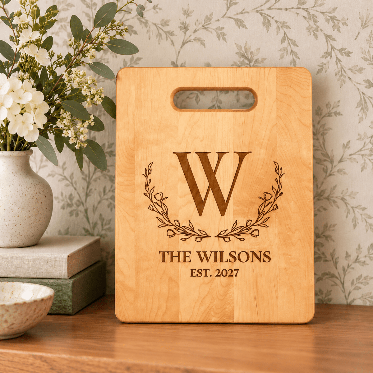 Personalized maple cutting board engraved with Wilson family name, floral wreath monogram, and established year, displayed with flowers and greenery.