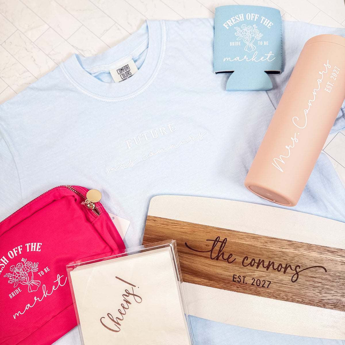 Example Mystery Bridal Box with personalized bride tee, tumbler, engraved board, koozie, pouch and napkins on a light blue shirt