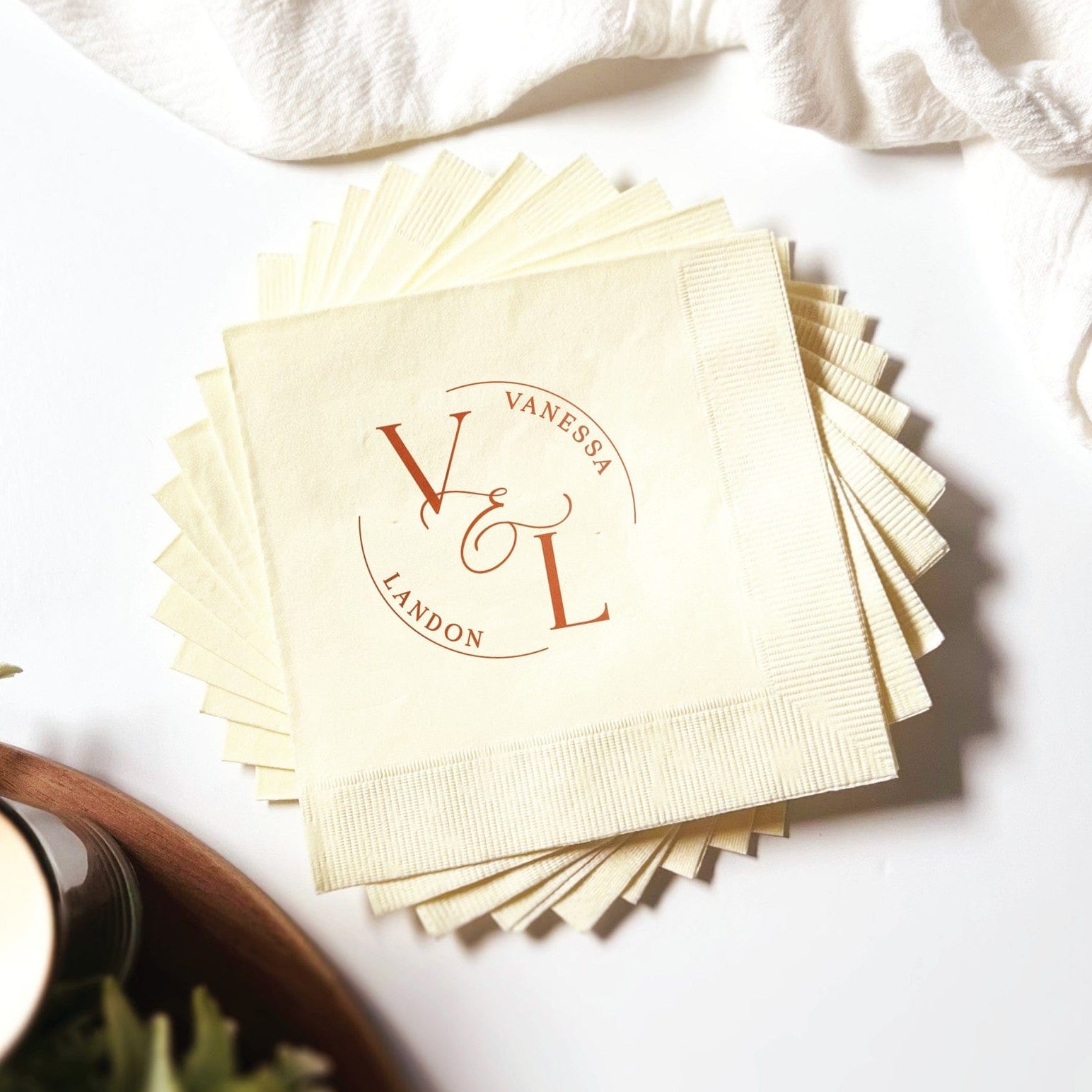 A stack of off-white personalized paper cocktail napkins featuring a reddish-brown monogram with large intertwined initials and text within an elegant oval design, suitable for a wedding or special event.