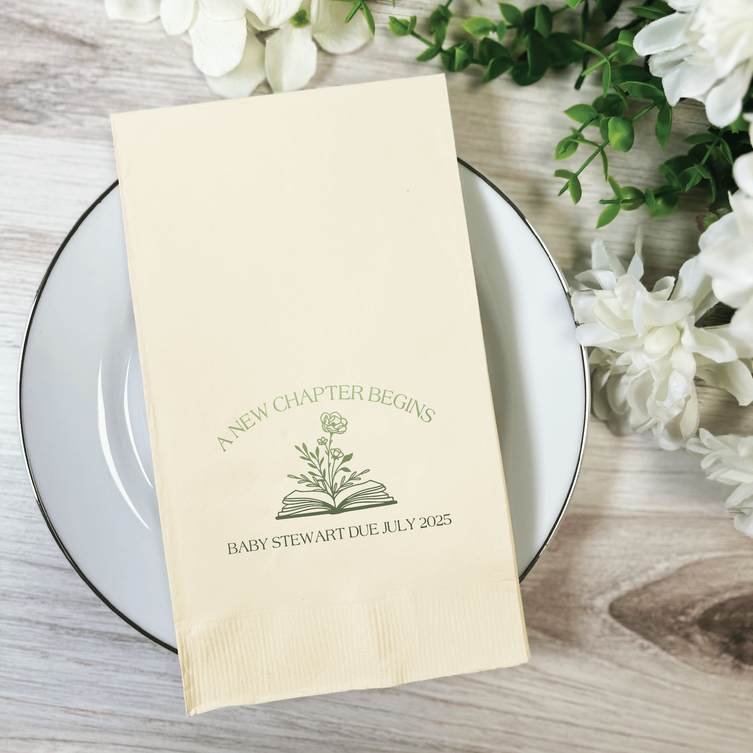 Cream napkin with green foil-stamped book and flower design, personalized with "A New Chapter Begins" and baby shower date, displayed on white plate.