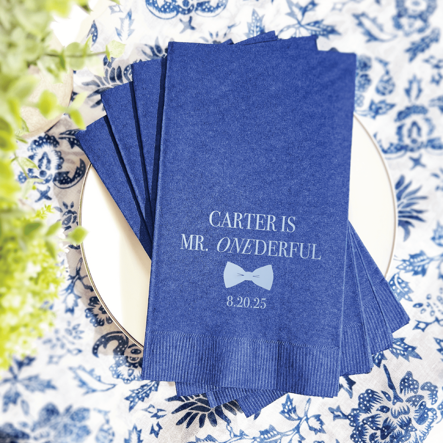 Stack of blue napkins with "Carter is Mr. Onederful" and date "8.20.25" in white foil stamping, arranged on a decorative blue and white plate.