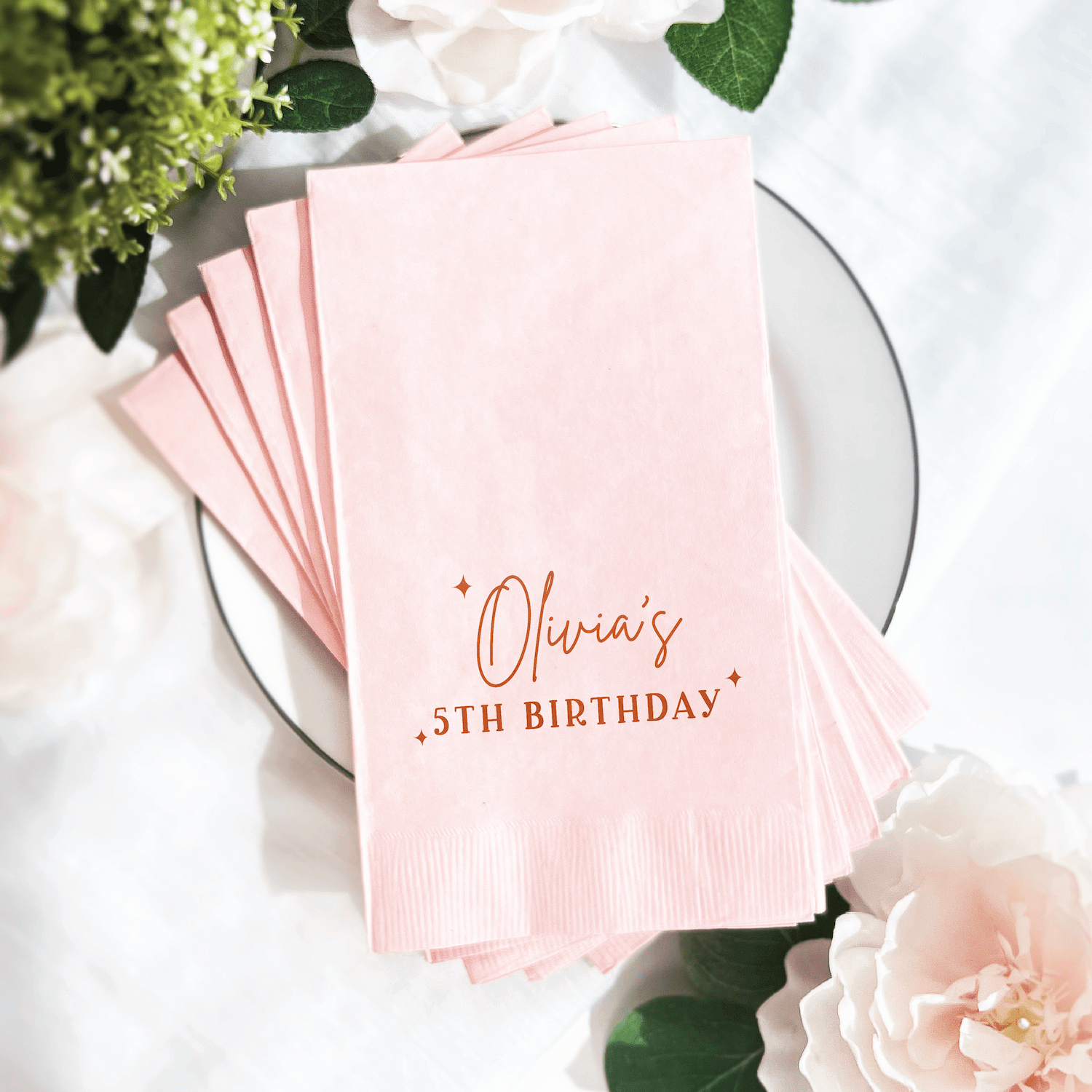 Stack of pink napkins with "Olivia's 5th Birthday" in rose gold foil lettering, displayed on a white plate with flowers.