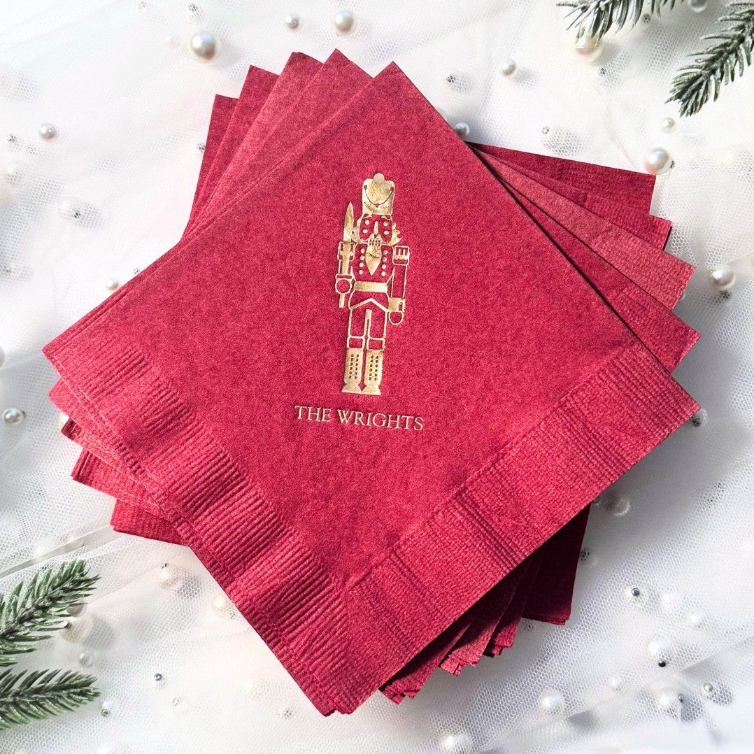 Red paper napkins featuring a gold foil Nutcracker design, surrounded by festive holiday greenery and pearl accents, suitable for a Christmas celebration.