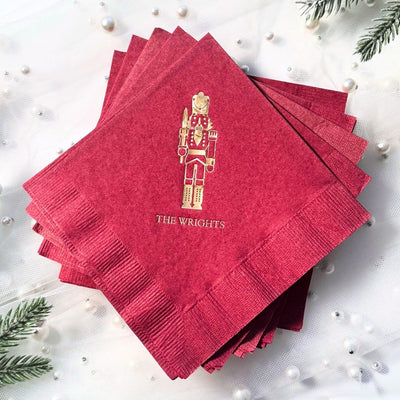 Red paper napkins featuring a gold foil Nutcracker design, surrounded by festive holiday greenery and pearl accents, suitable for a Christmas celebration.