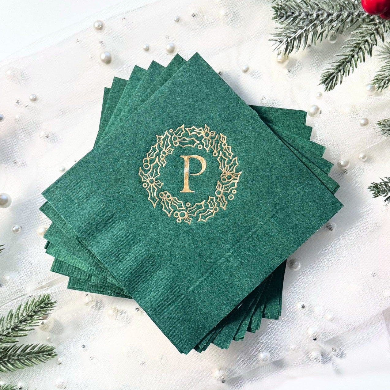 A stack of dark green paper napkins featuring a gold foil holly wreath design with a centered initial, perfect for a Christmas or winter event.