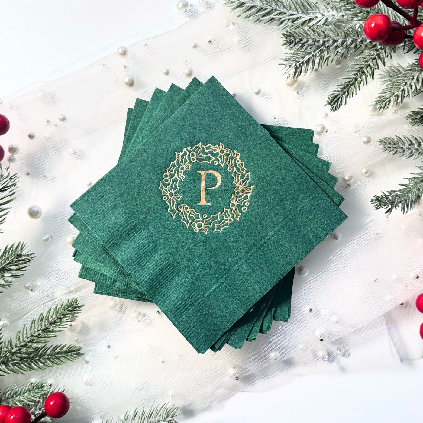 A stack of dark green paper napkins featuring a gold foil holly wreath design with a centered initial, perfect for a Christmas or winter event.