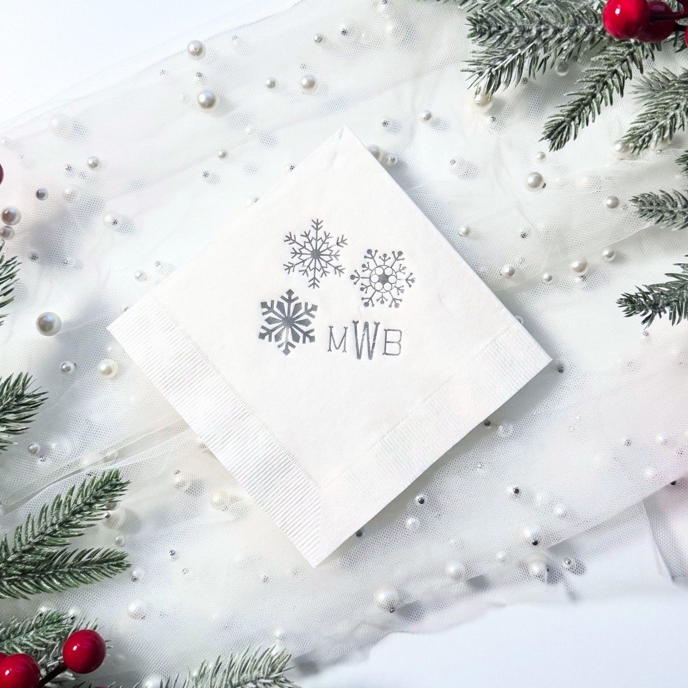 A white square paper cocktail napkin featuring a trio of silver snowflake designs, perfect for a winter wedding or holiday celebration.