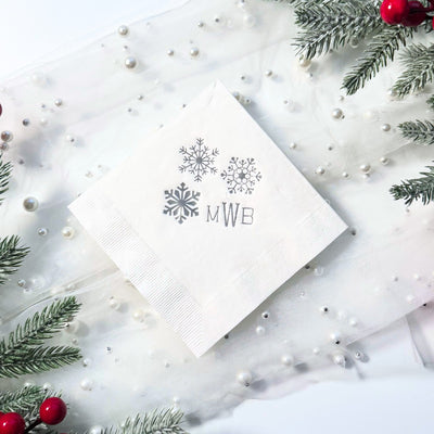 A white square paper cocktail napkin featuring a trio of silver snowflake designs, perfect for a winter wedding or holiday celebration.