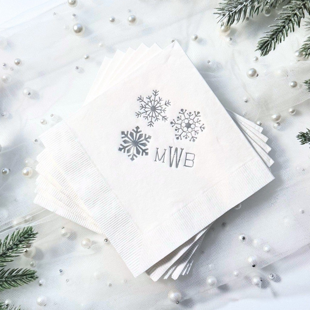A stack of white paper napkins with a subtle ribbed texture, featuring three metallic silver snowflake designs, are arranged on a sheer white fabric adorned with pearls and surrounded by frosted pine branches and red berries, suitable for a winter event.