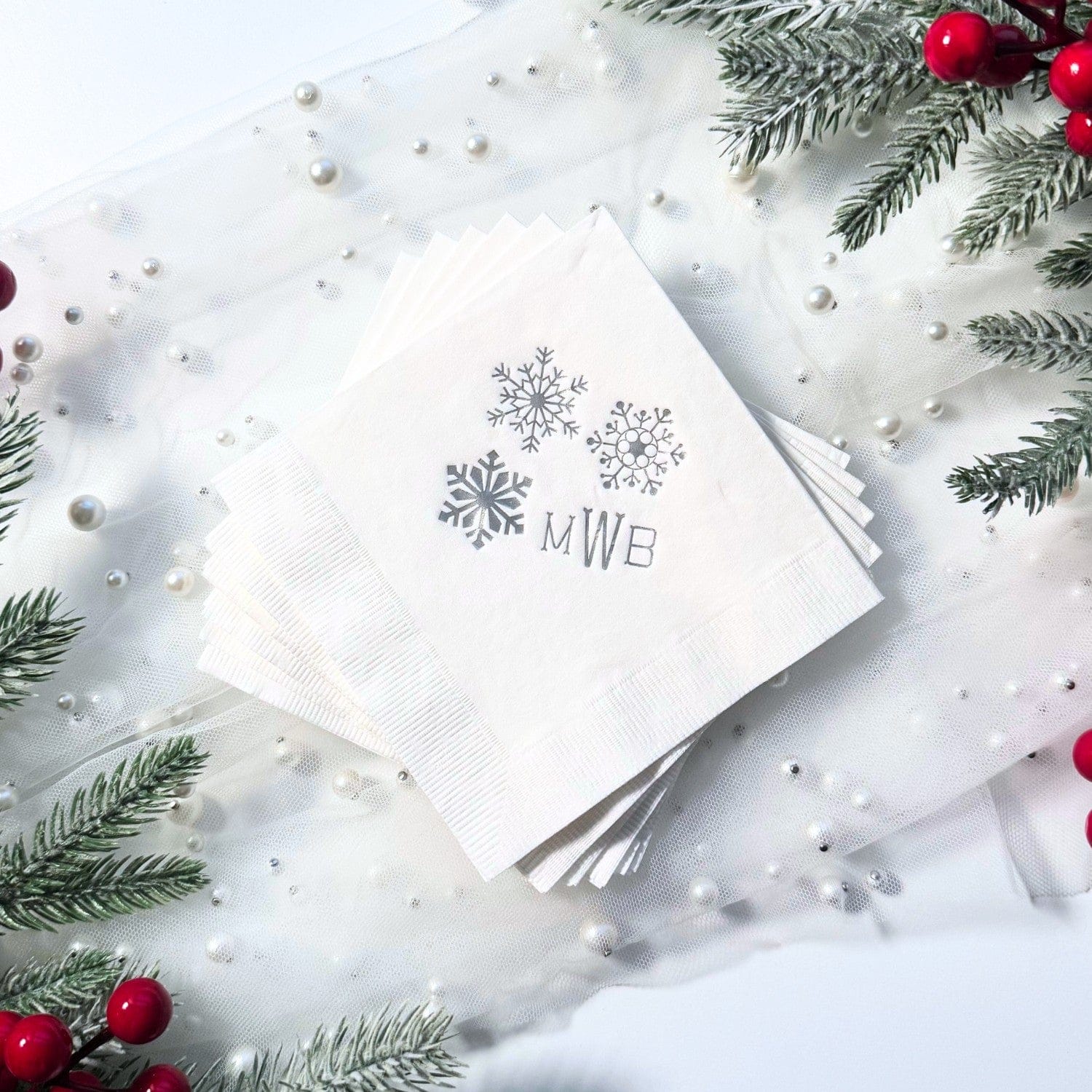 A stack of white paper napkins with a subtle ribbed texture, featuring three metallic silver snowflake designs, are arranged on a sheer white fabric adorned with pearls and surrounded by frosted pine branches and red berries, suitable for a winter event.