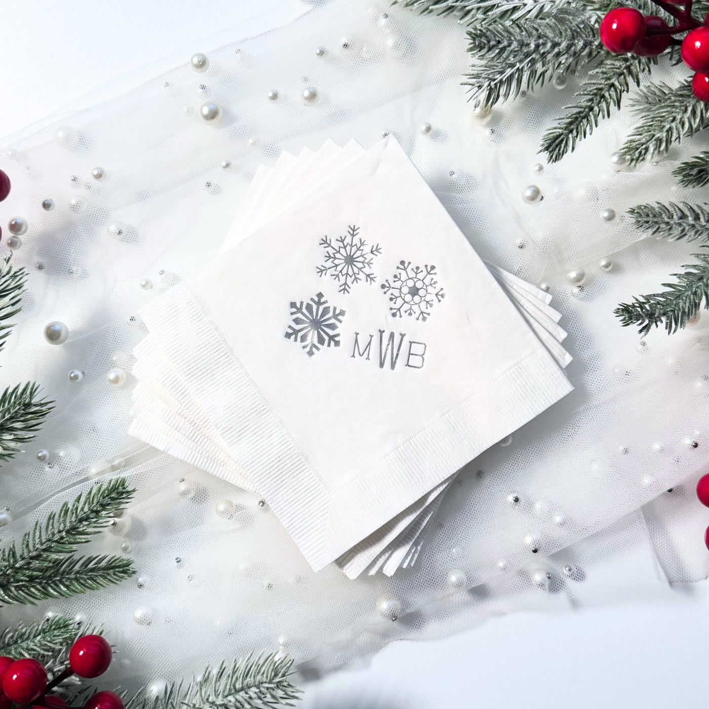 A stack of white paper napkins with a subtle ribbed texture, featuring three metallic silver snowflake designs, are arranged on a sheer white fabric adorned with pearls and surrounded by frosted pine branches and red berries, suitable for a winter event.