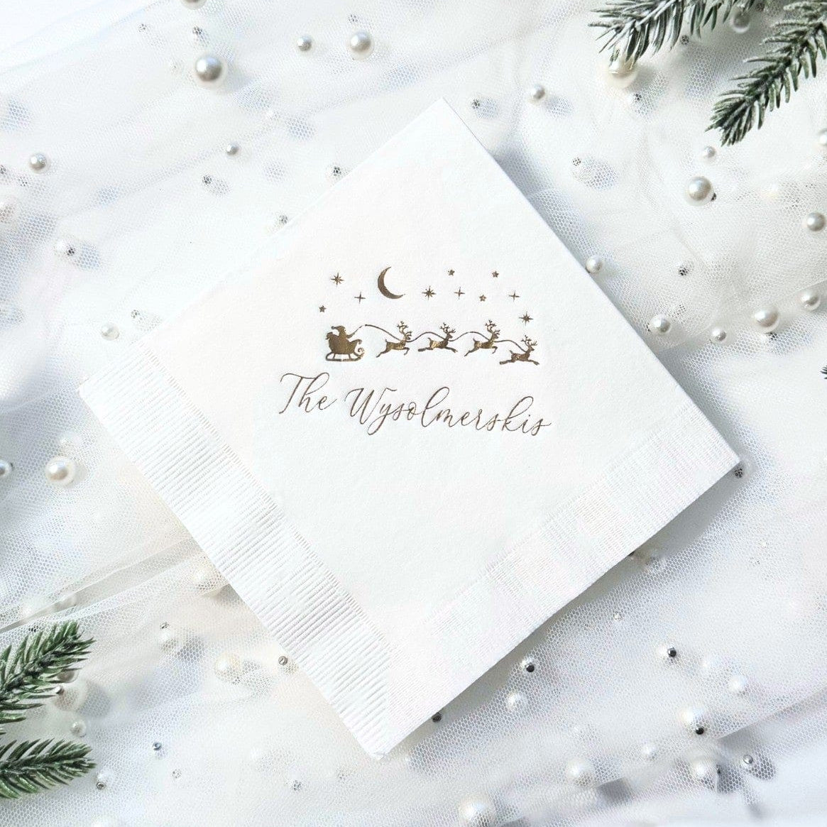 A white square paper napkin featuring a gold foil illustration of Santa's sleigh and reindeer under a crescent moon and stars, suitable for a Christmas or holiday event.