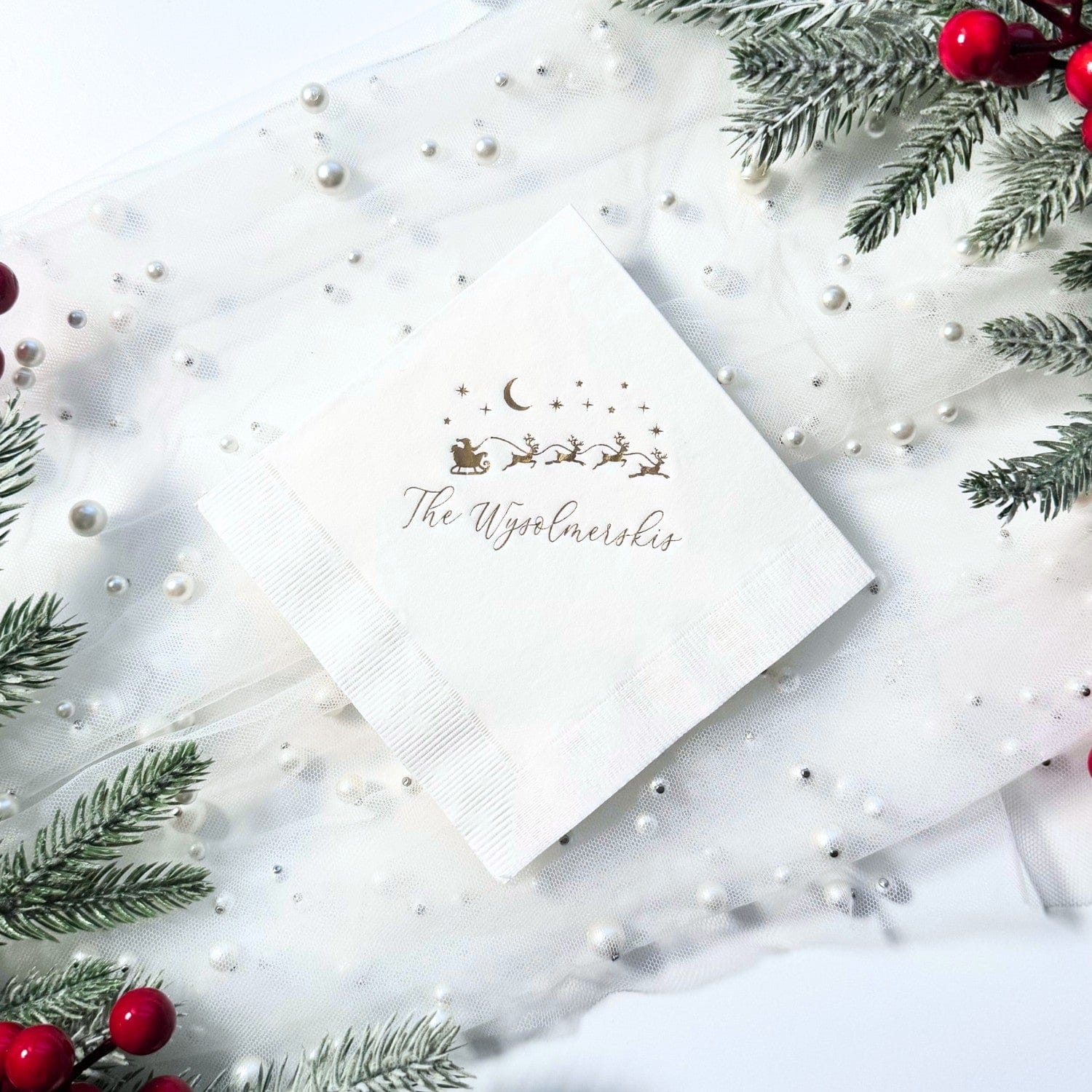 A white square paper napkin featuring a gold foil illustration of Santa's sleigh and reindeer under a crescent moon and stars, suitable for a Christmas or holiday event.