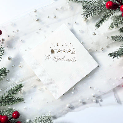 A white square paper napkin featuring a gold foil illustration of Santa's sleigh and reindeer under a crescent moon and stars, suitable for a Christmas or holiday event.