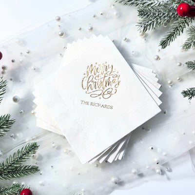 A stack of white paper cocktail napkins featuring festive gold foil lettering, ideal for a Christmas celebration.