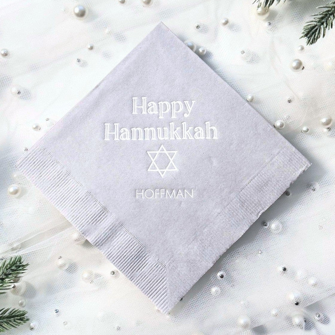 A light grey personalized paper napkin with a subtle embossed design is elegantly presented on a white sheer fabric adorned with pearls and frosted evergreen branches, suitable for a holiday celebration.