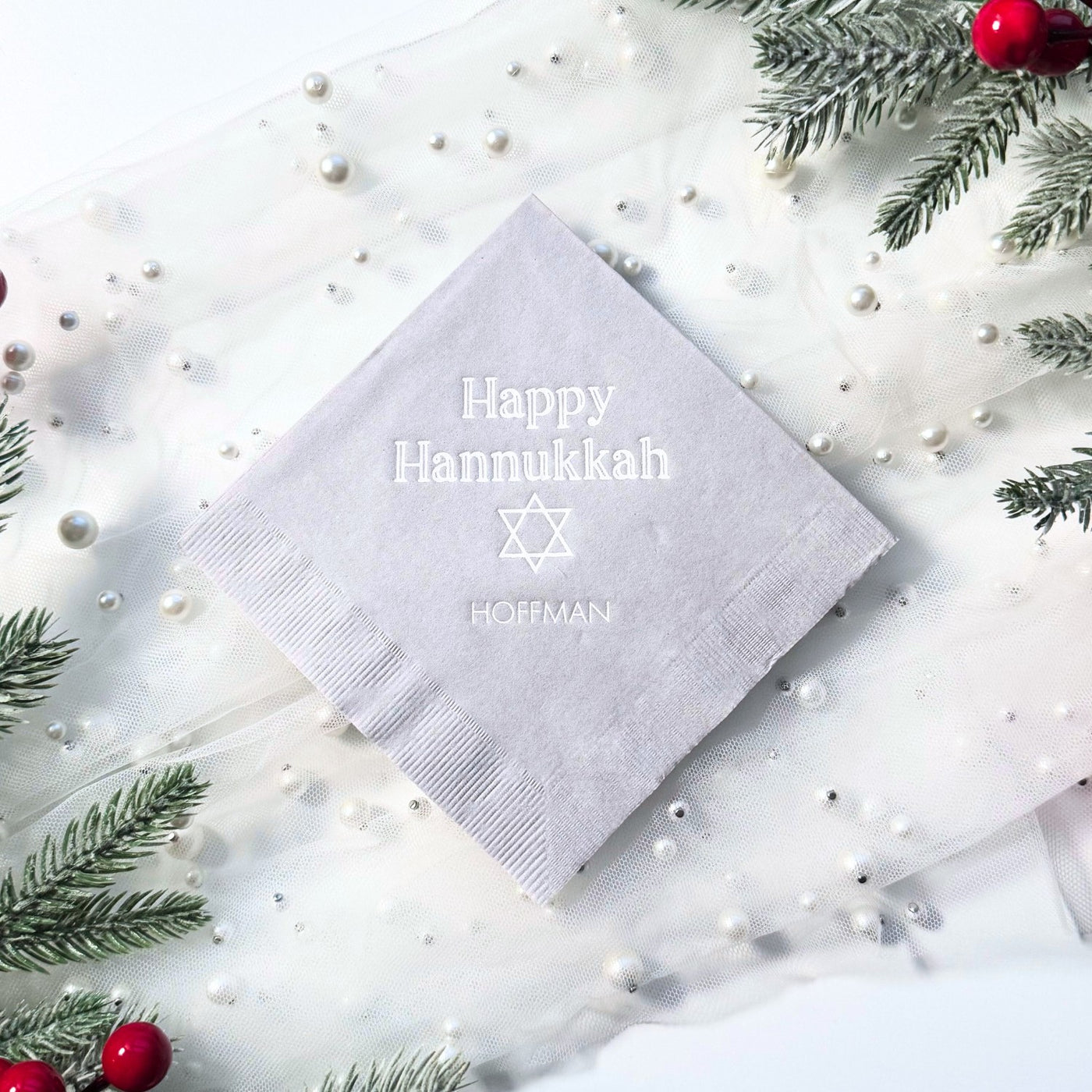 A light grey personalized paper napkin with a subtle embossed design is elegantly presented on a white sheer fabric adorned with pearls and frosted evergreen branches, suitable for a holiday celebration.