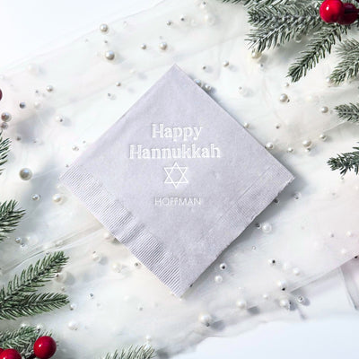 A light grey personalized paper napkin with a subtle embossed design is elegantly presented on a white sheer fabric adorned with pearls and frosted evergreen branches, suitable for a holiday celebration.