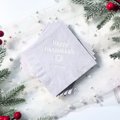 A stack of light gray paper napkins featuring an embossed holiday greeting and a religious symbol, perfect for a Hannukkah celebration.