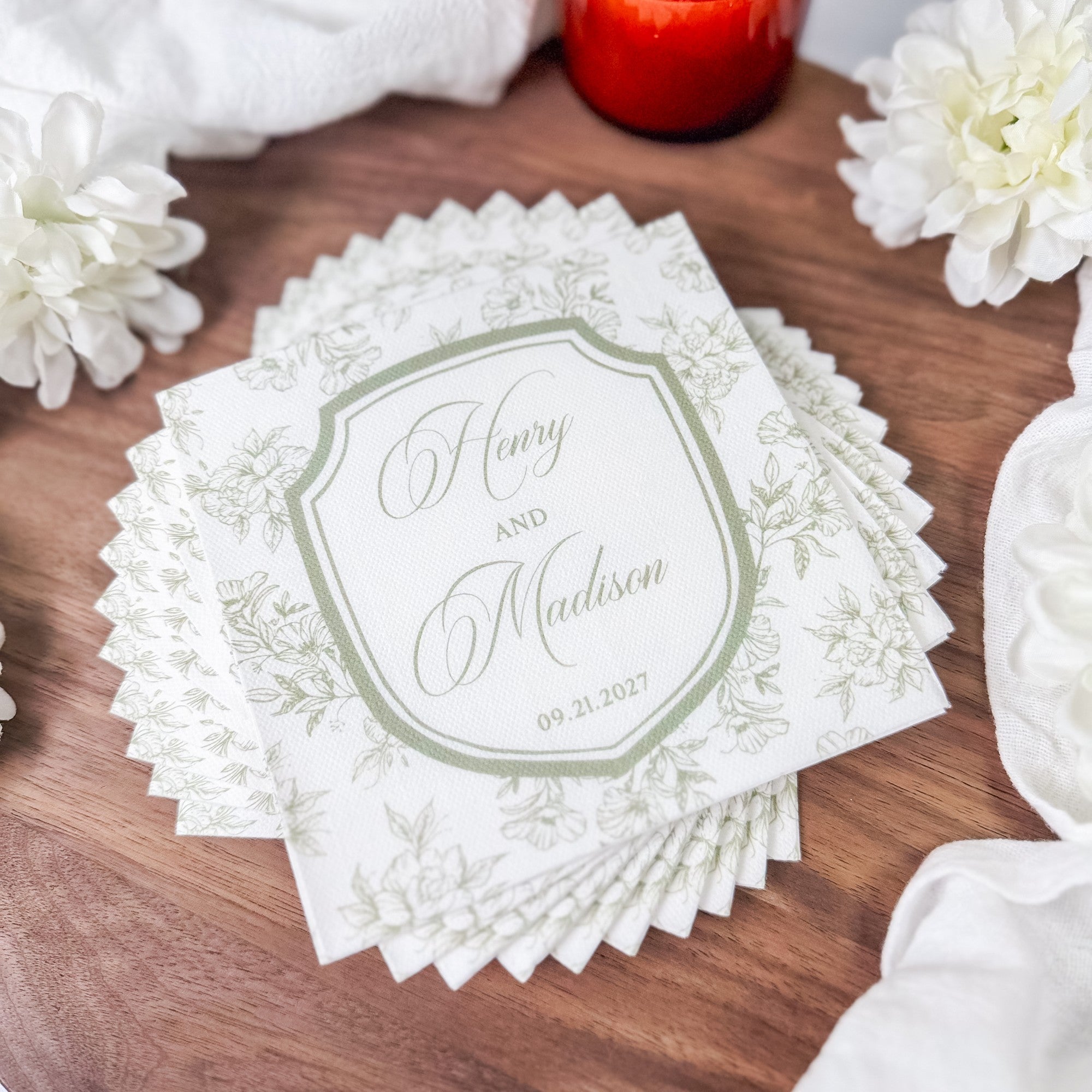 A stack of white paper napkins with scalloped edges features an elegant green floral pattern and a central shield design, ideal for wedding or event decor.