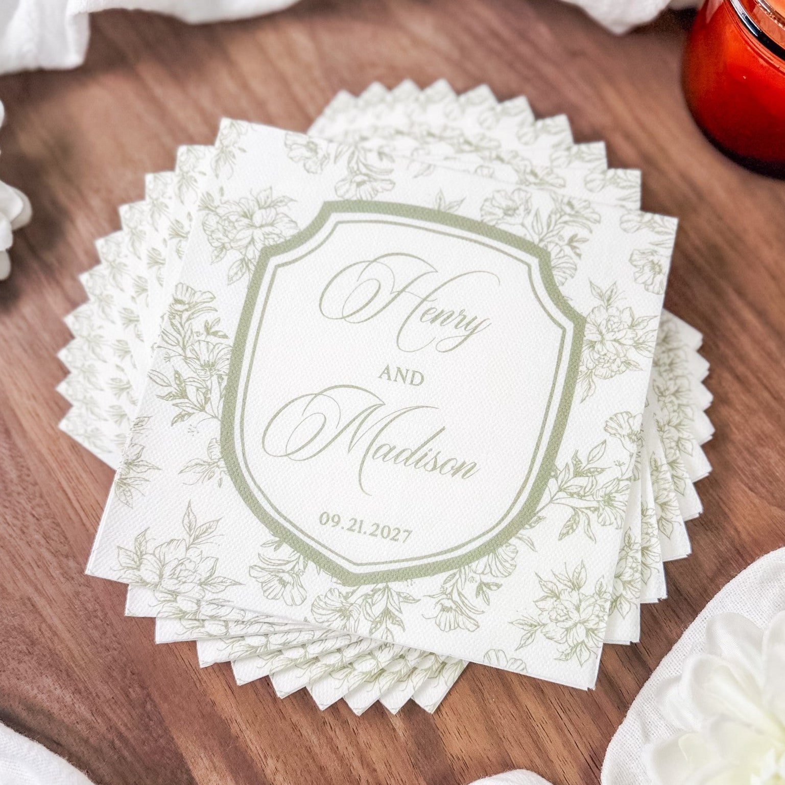 A stack of white personalized paper wedding napkins featuring a light green floral design around a central crest.