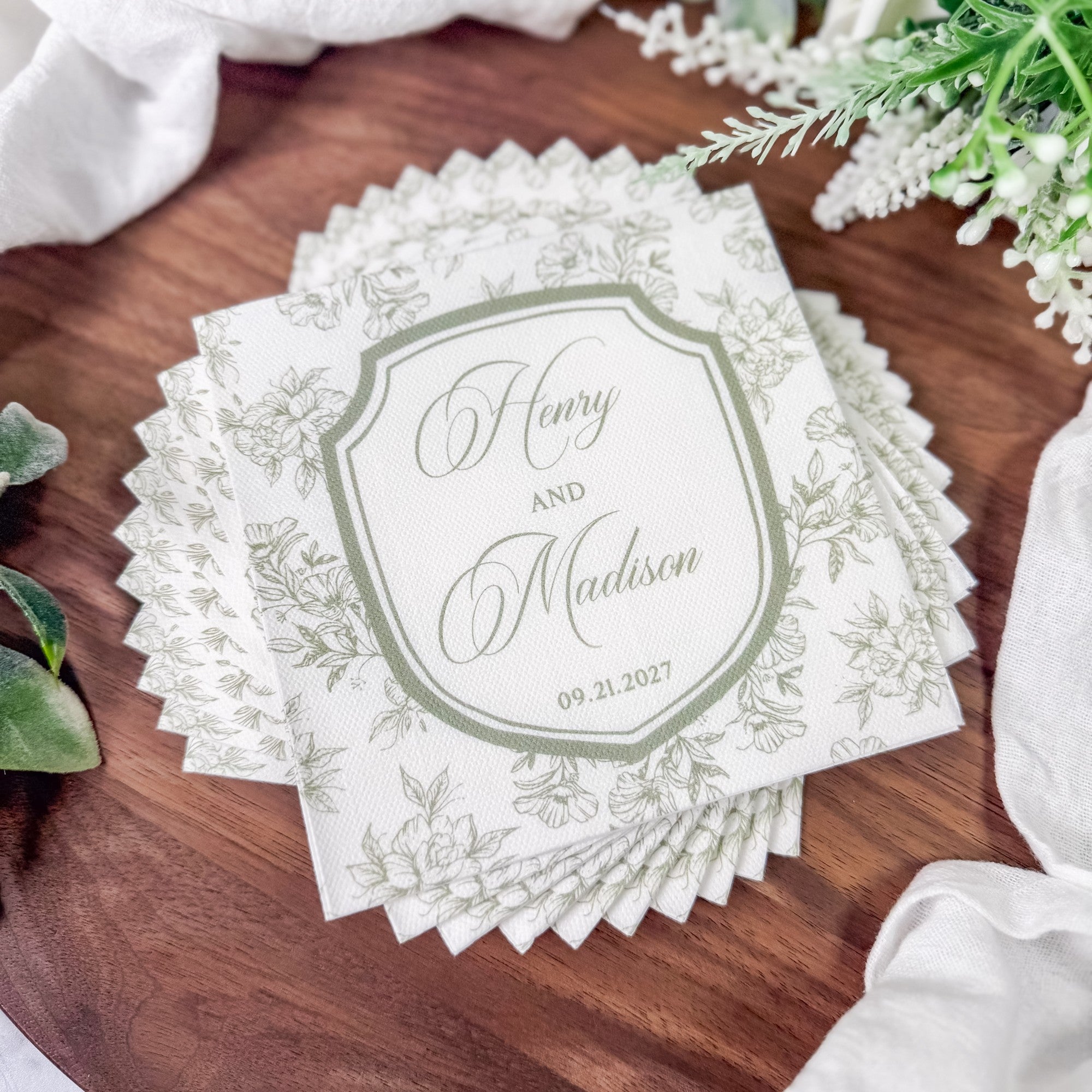 A stack of personalized white paper cocktail napkins featuring a delicate green floral pattern, a central crest design, and elegant scalloped edges, perfect for a wedding.