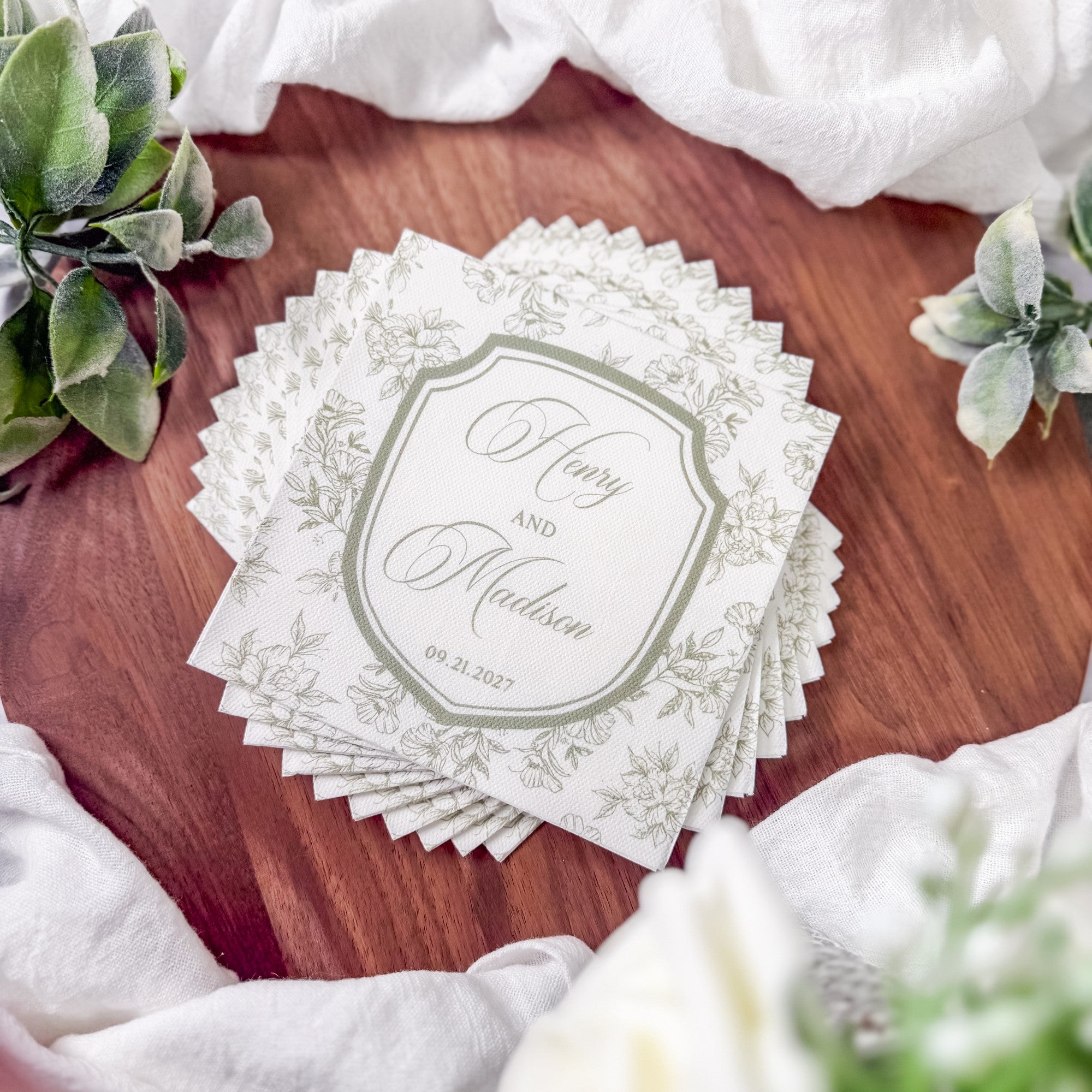 A stack of white square paper napkins with scalloped edges, featuring a delicate light green floral pattern and a personalized crest design, ideal for wedding or elegant event decor.