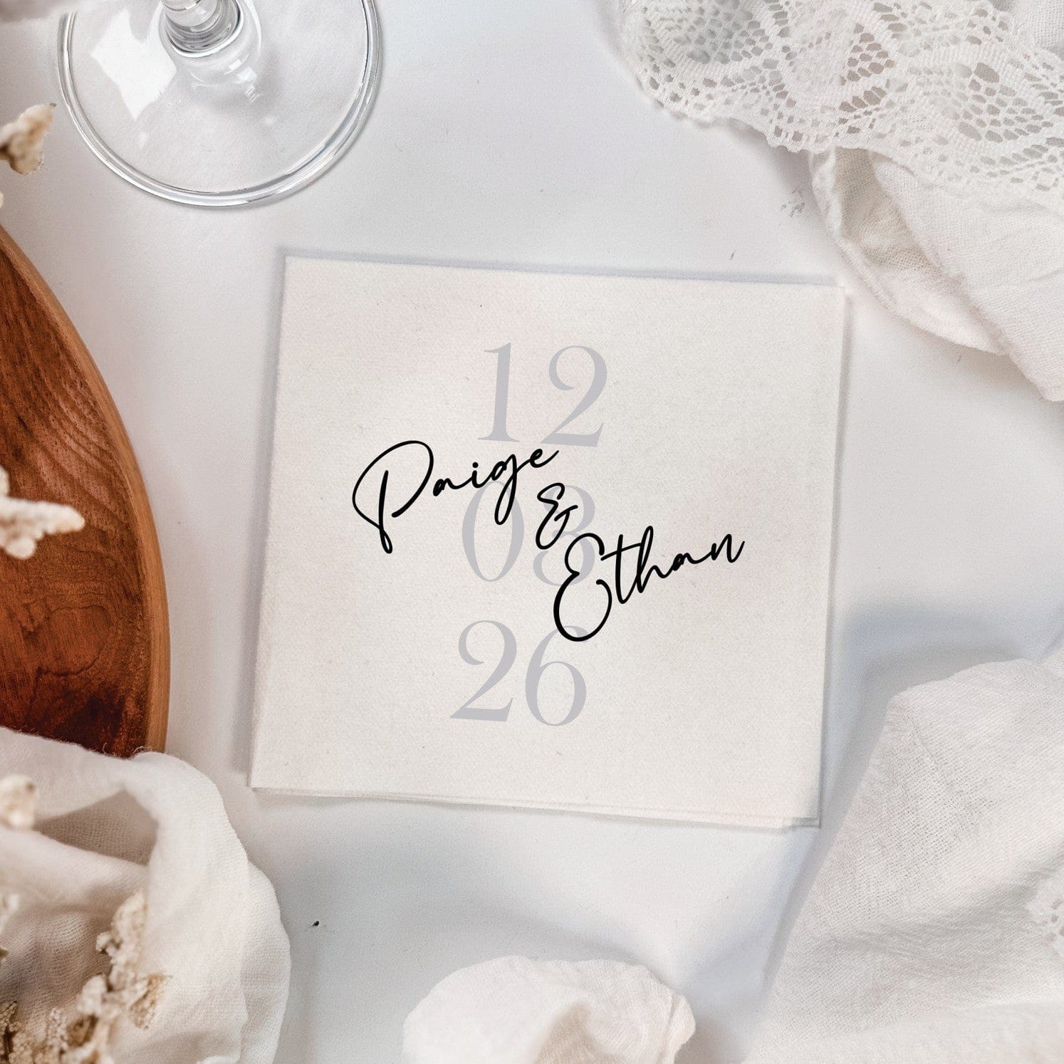 A personalized light-colored paper napkin featuring elegant black script and subtle gray numbers, perfect for a wedding or special event, displayed amongst white draped fabric and dried floral accents.