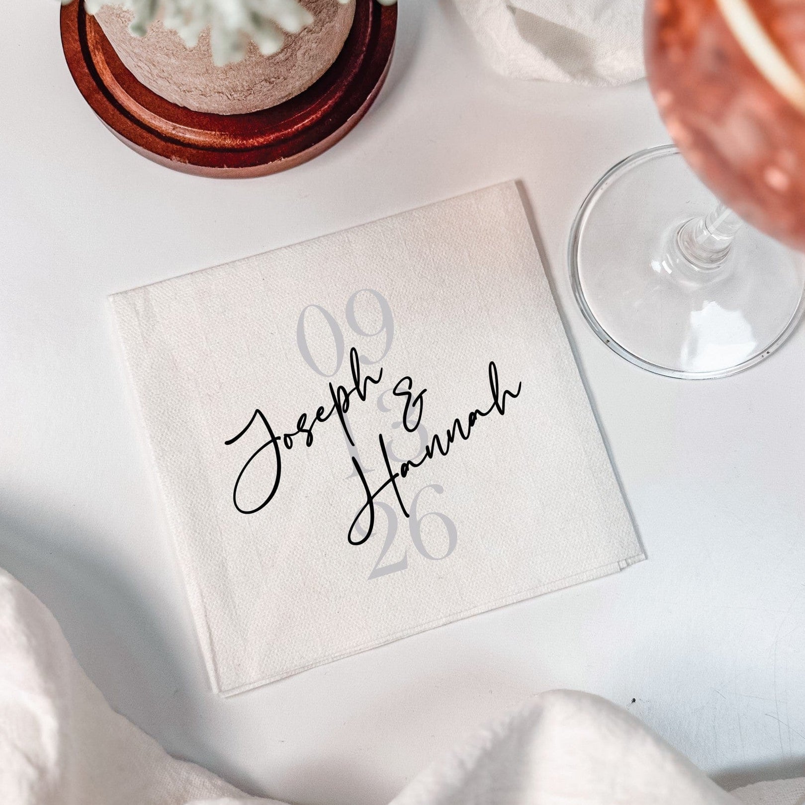 A white square disposable wedding napkin featuring a custom black script design and subtle gray numbers, styled on a white surface with draped fabric and a drink, for event decor.