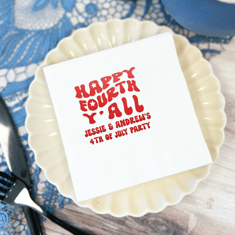 White foil-stamped napkin with red "Happy Fourth Y'all" text and personalized names, placed on cream scalloped plate with blue patterned tablecloth.