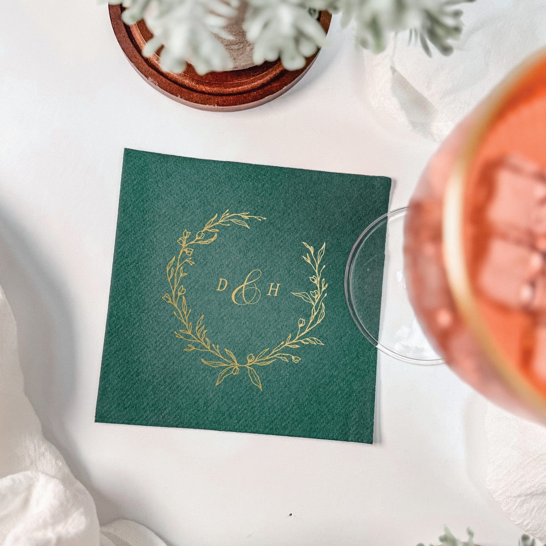 A square, dark green paper napkin featuring a gold foil floral wreath design, suitable for a wedding or elegant event.