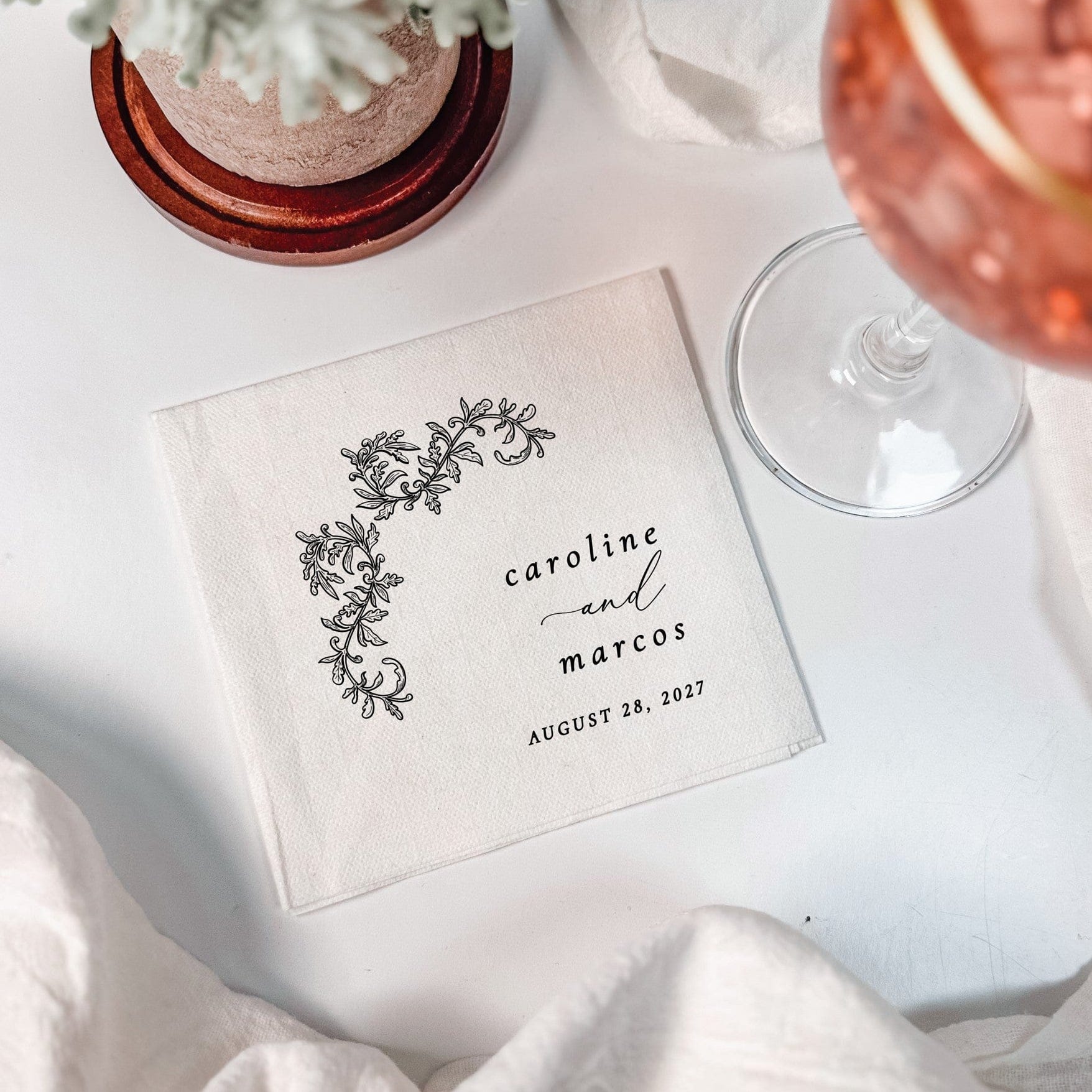 A square white linen napkin, personalized with a black floral and leafy design, serves as an elegant table accessory for a wedding celebration.