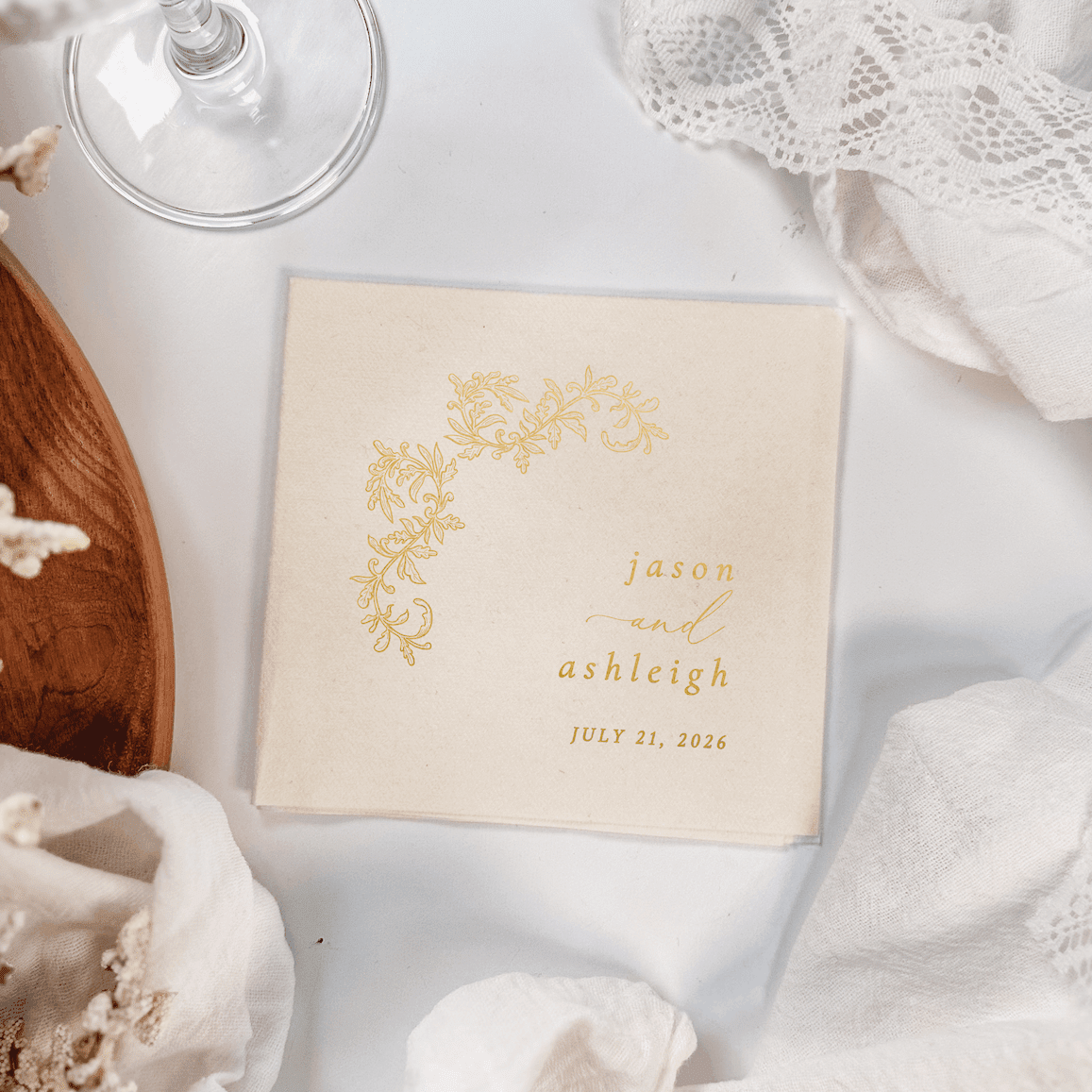 Cream cocktail napkin with gold foil floral corner design and personalized names "jason and ashleigh" with wedding date July 21, 2024.