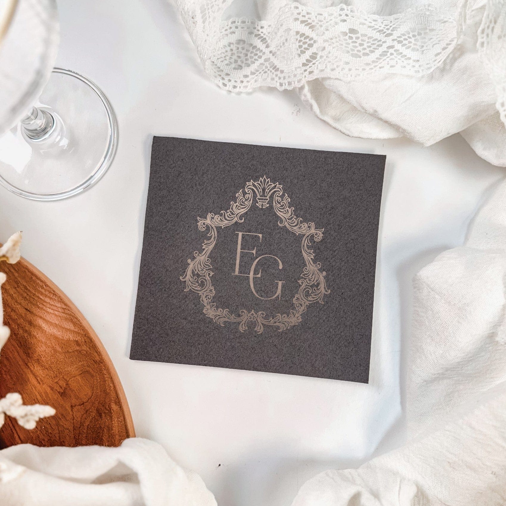 A dark gray square linen or paper napkin features an elegant light-colored monogram within an ornate frame, perfect for a sophisticated wedding or event.