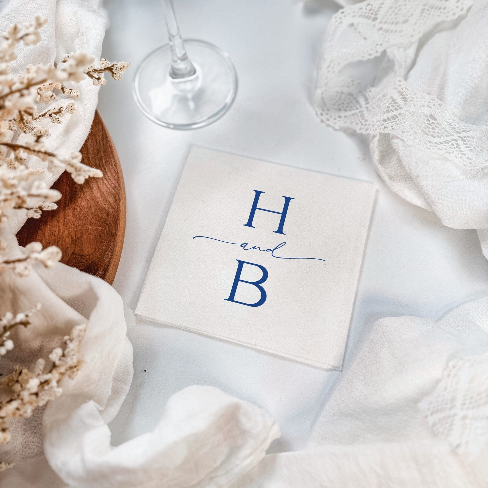 A stack of elegant white linen-like wedding napkins featuring a custom blue initial design, artfully arranged with delicate white fabric and dried floral accents.