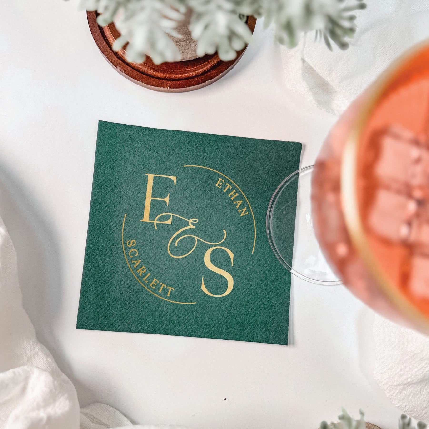 A dark green square linen-feel napkin features a personalized gold foil printed circular monogram design, perfect for a wedding or elegant event.