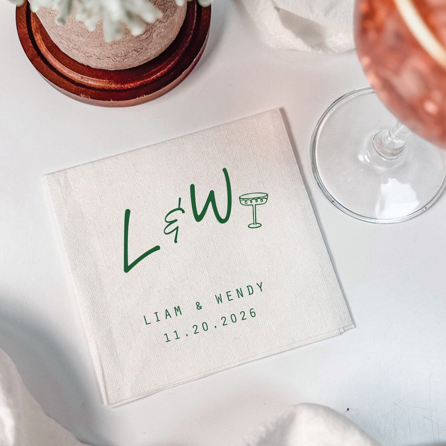 A white personalized linen-feel cocktail napkin, perfect for a wedding or event, features a custom initial and drink icon design printed in dark green.